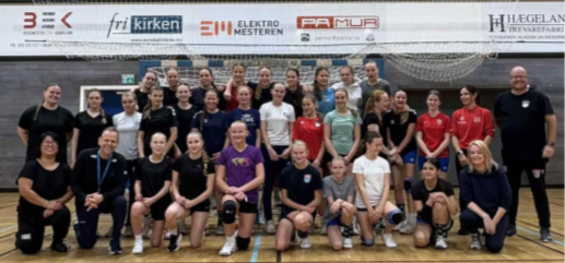 Gruppebilde av spillere og trenere i sportsklær på en innendørs bane, med sponsorbanner i bakgrunnen. Bildet er tatt etter treningsøkten med Thorir Hergeirsson.