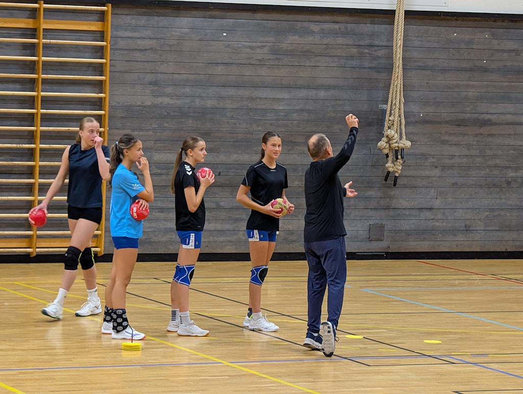 Trener Thorir Hergeirsson instruerer fire unge håndballspillere i en gymsal. Spillerne står på rekke med røde baller, mens treneren gestikulerer foran dem. I bakgrunnen sees trevegger, klatretau og ribbevegg.